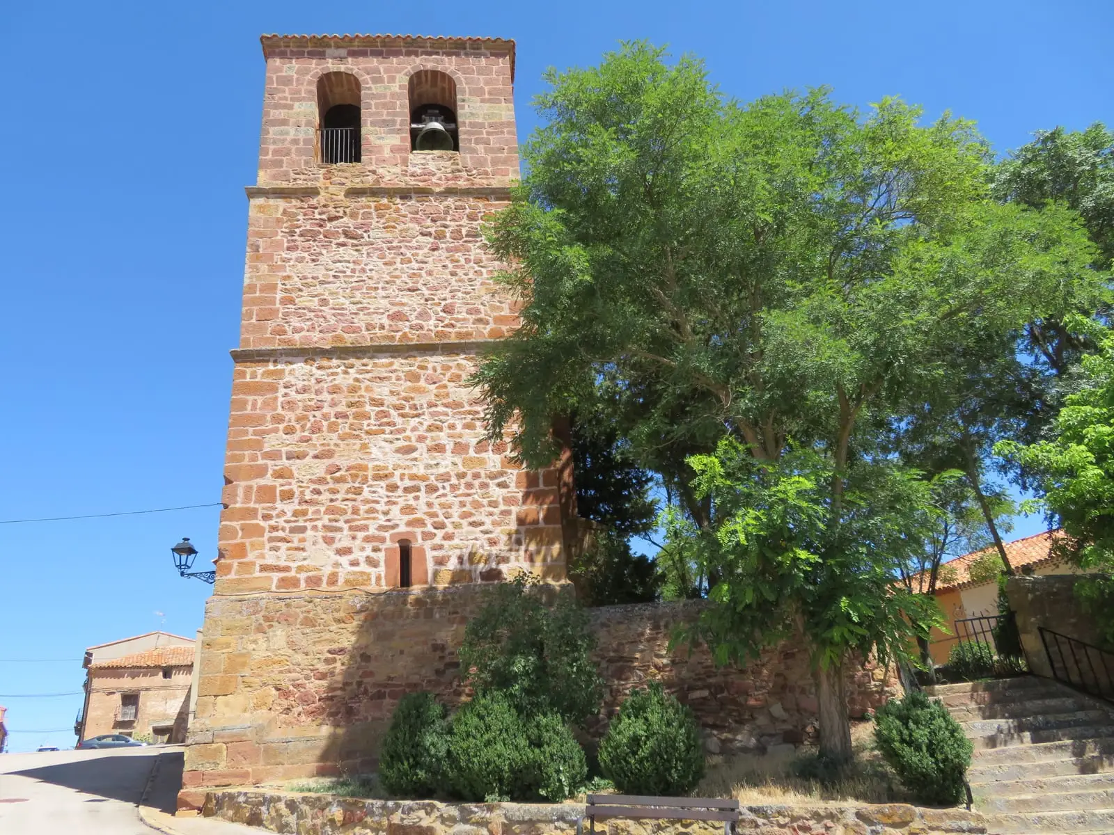 Reddish stone bell tower in El Pobo de Dueñas, beside a tree and village houses.