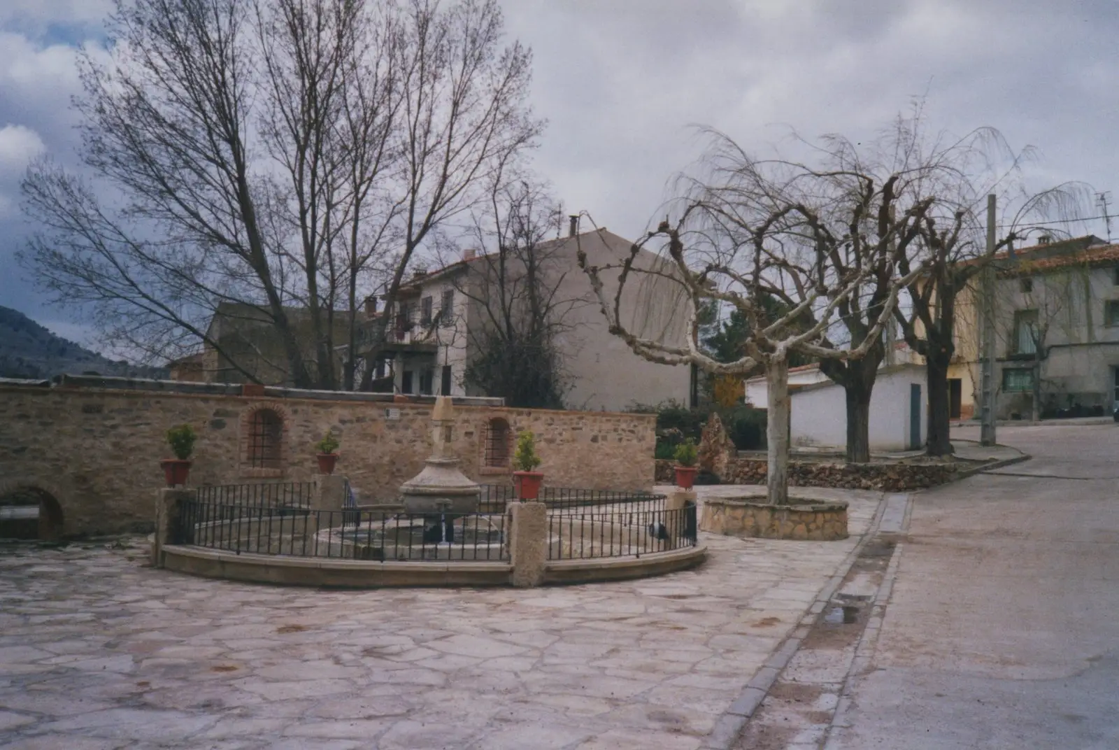 Fuentelsaz square with a stone fountain, leafless trees and surrounding houses.